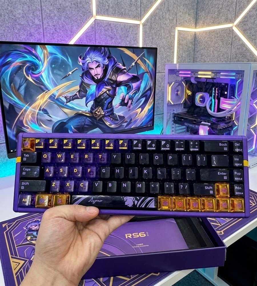 Hand holding the purple and black ATK RS6 Ultra keyboard with amber accent keys above its retail box in front of a gaming monitor