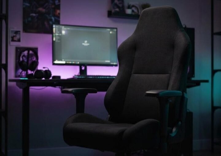 Black fabric gaming chair with a wide seat in front of a desk and monitor lit by purple and teal lighting
