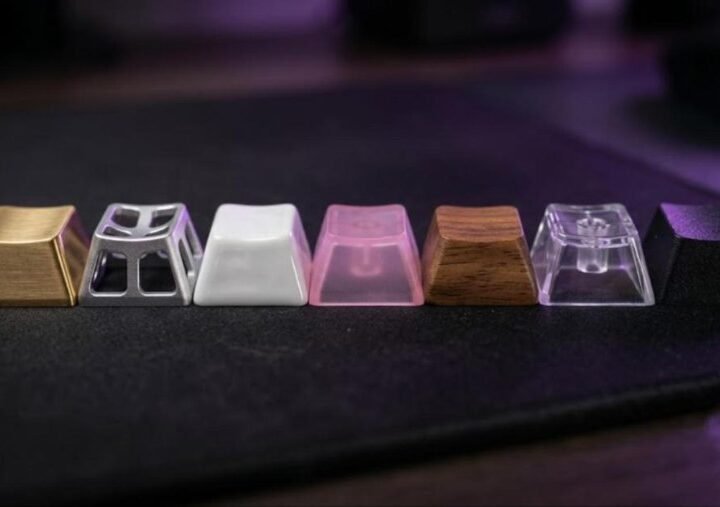 Seven keycap materials side by side: brushed gold metal, hollow aluminum, white ceramic, pink jelly polycarbonate, walnut wood, clear polycarbonate and black PBT